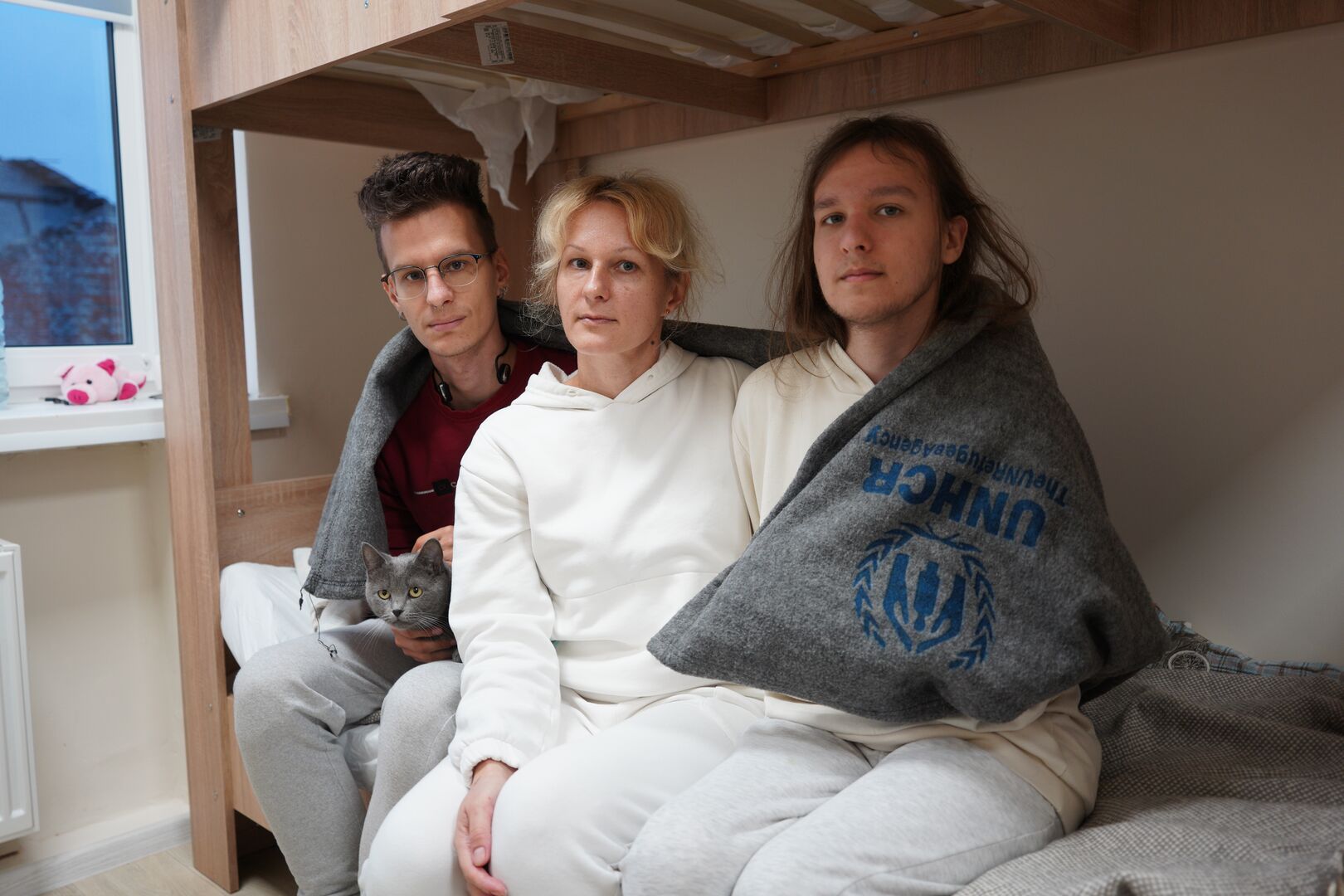 A mother and her two adult sons sit on a bunk bed wrapped in a UNHCR blanket