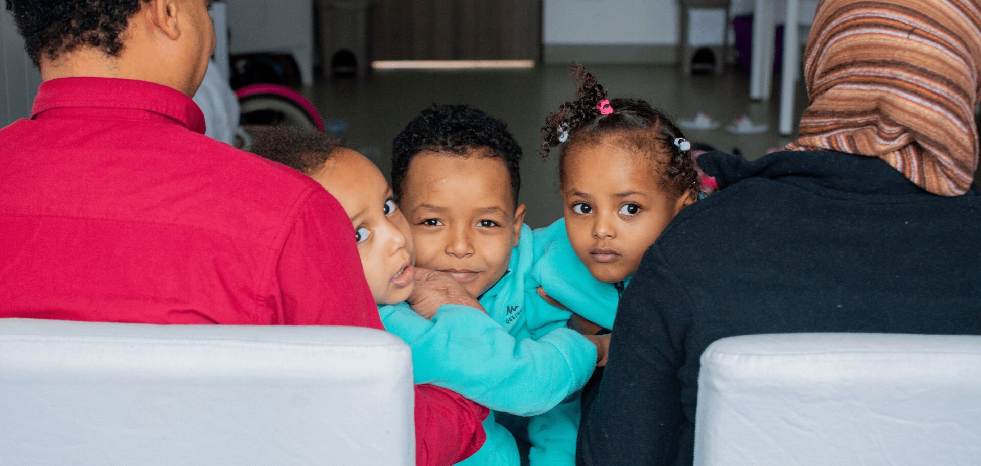 Three Eritrean children peek at the camera from their parents' laps